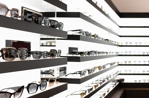 Shelves in an eyeglass store filled with stacked eyeglass frames on display in rows, partially blurred.