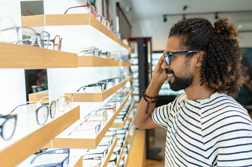 Multiracial long-haired modern Arab man in glasses store, examining and trying on and buying reading glasses and sunglasses.