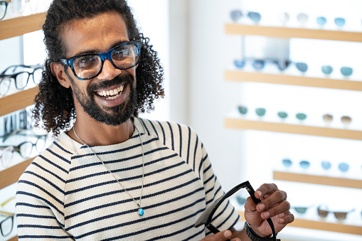 Multiracial long-haired modern Arab man in glasses store, examining and trying on and buying reading glasses and sunglasses.