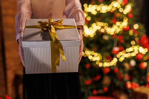 Close up shot of unrecognizable young woman standing by the glistening Christmas tree and hanging you a silver colored gift box with a gold colored bow on top.