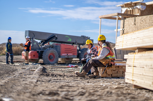 A small group of construction workers sit on a stack of lumber as they take a quick lunch break.  They are each wearing proper safety gear and are talking amongst themselves as they sit onsite.