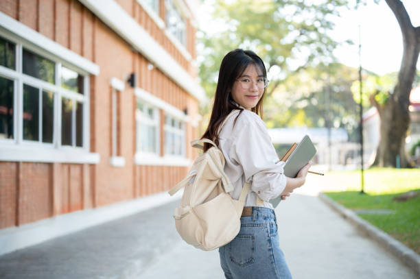 istock Young beautiful Asian female college student with a backpack and laptop computer is in a campus. 1839172476