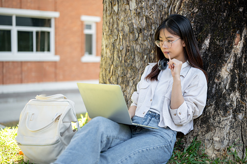 A positive and thoughtful Asian female student is using her laptop computer under the tree in a campus park, doing homework online.