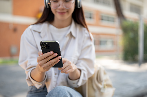 A pretty and positive Asian female student is using her smartphone and listening to music on her headphones while relaxing on a bench in a campus park. University life, leisure, lifestyle