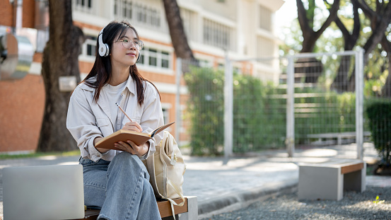A positive young Asian female in casual clothes is listening to music on her headphones and keeping her diary while resting on a bench in a park. student, freelancer, leisure and lifestyle