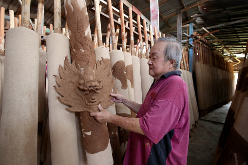 An Asian senior man is holding oversized dragon design Joss Stick sculpture in Factory - Daily Production Activities