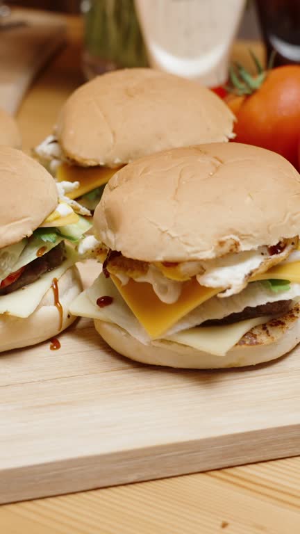 Vertical video. Homemade Burgers on a Wooden Table, with ingredients placed nearby along with sauce bottles. Cinematic lighting and camera movements.