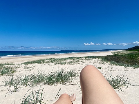 Horizontal view of looking at legs in foreground laying on sand dunes with grassy coverage looking out to clear blue sky and ocean waters on hot summer day at Brunswick Heads beach NSW Australia