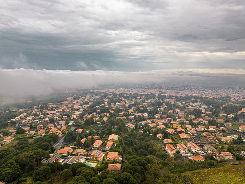 Aerial view of Nicolosi, Italy, showcasing red-roofed homes against a dramatic stormy sky, highlighting the tranquil yet vibrant Sicilian landscape.