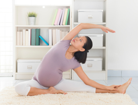 Pregnancy yoga meditation. Full length healthy 8 months pregnant calm Asian woman meditating or doing yoga exercise at home. Relaxation yoga sitting side stretch positions.