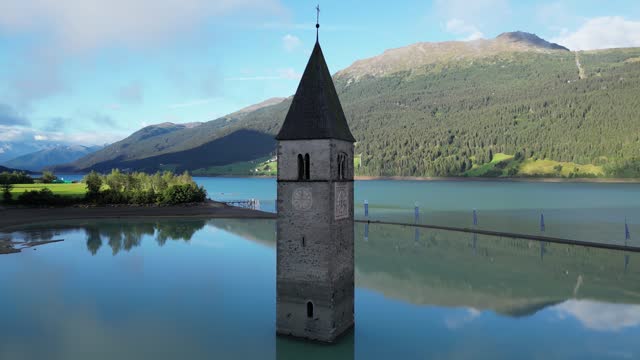 Orbit around steeple of submerged church in lake Reschensee and mountain alps panorama in South Tyrol, Italy
