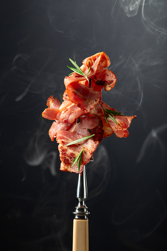 Hot fried bacon slices with rosemary on a fork. Copy space.