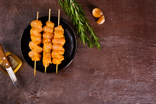 Raw chicken skewers in marinade with spices on a black plate and on a brown table