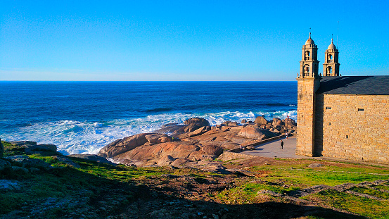 Virxe da Barca Sanctuary church, bell towers, coastline and Atlantic ocean view, breaking waves, horizon, clear sky. Muxía, Costa da Morte, Galicia, Spain