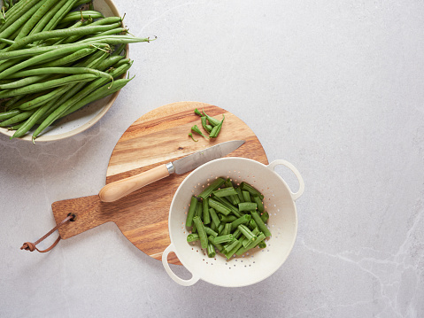 Fresh raw green bean ends on a wooden cutting board next to a knife, cut chunks in a colander and whole pods in a bowl. Vegetarian food preparation. Top view on gray kitchen countertop.