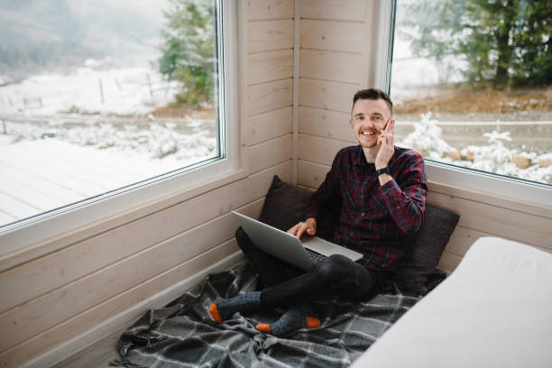istock Man talking on phone. Remote work from cozy place. Male remote working on laptop in front of panoramic window with a great view of mountains. Workplace in house in snowy morning. Freelance lifestyle 1839576814