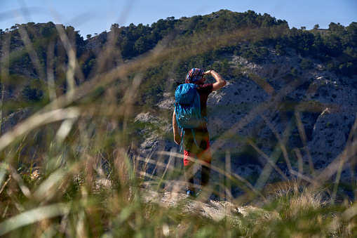 Full body of male hiker with backpack standing on grassy mountain slope and looking away on sunny day