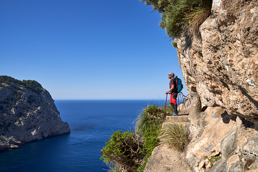 Side view of man with backpack standing on stone footpath on cliff and observing rippling ocean during summer vacation