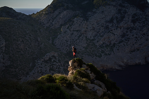 Anonymous male hiker standing on rough rocky edge and admiring picturesque landscape with mountains at sunset