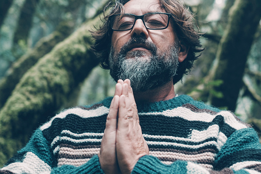 Mature bearded man portrait meditate in the middle of green environment beautiful forest. Aged male with eyewear meditating in the outdoors nature park with closed eyes and hands clasped. Serene life