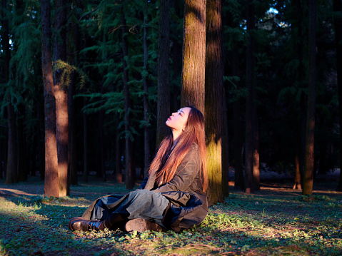 Portrait of elegant Chinese girl in black coat sit on meadows enjoy carefree time in forest park in sunny day. Outdoor fashion portrait of glamour young Chinese cheerful stylish woman.