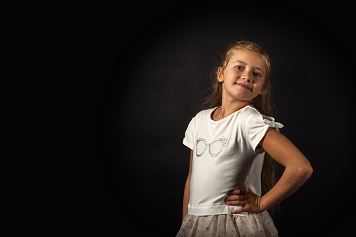 Portrait of little girl 7-8 year old in white attire posing at black backdrop, smile looking at camera. Pensive lovely child lady expressing emotions, studio shot. Youth concept. Copy ad text space