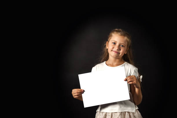 istock Cute child girl holding white empty space blank, poses at black backdrop, smiling looking at camera. Kid model promote mockup template, blank paper in hands. Advertising concept. Copy ad text space 1839716428