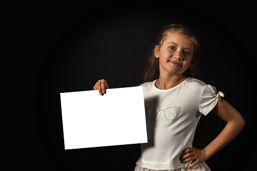 Cute kid girl model holding white empty space blank paper in hand at black, looking at camera. Adorable child promote mockup template,  studio shot in shadow. Advertising concept. Copy ad text space