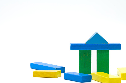 Two green wooden blocks standing isolated on a white background with a blue crossbar and triangle as a roof. Colorful toy blocks lying around.