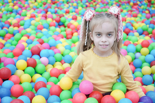 A beautiful blond girl with two ponytails sits among colorful bright balls. Close-up portrait. Cute happy girl in the playroom.