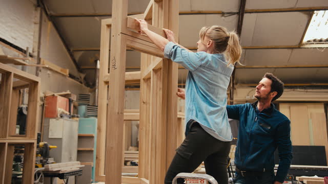 Male And Female Carpenters In Workshop Gluing Piece Of Wood To Window Frame