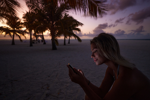 Young smiling woman reading a text message on smart phone during summer evening on the beach.
