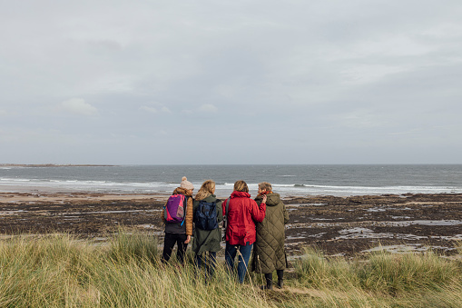 A rear view of a group of mature female friends who are stood looking out at the view in Bamburgh in the North East of England. They are having a mindful moment together. The women with the short blonde hair has recently beat breast cancer.