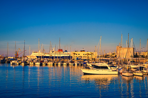 View of port area and bay in Heraklion, capital of Crete island, Greece. Ferries and cruises, fishing boats moored in harbor at sunset. Clear blue sky, deep blue waters, yachts, sailboats. Travel destination of Mediterranean sea, summer vacations.