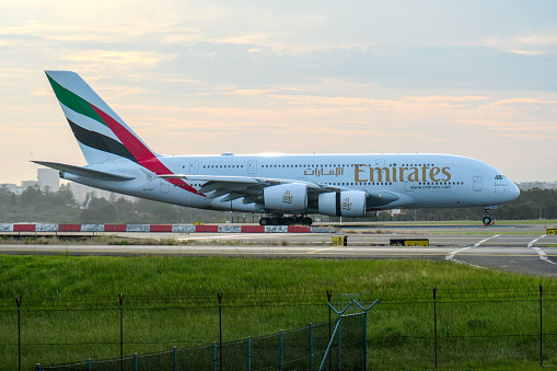An Emirates Airbus A380-842 plane, registration A6-EEK, landing from the south on the main runway of Sydney Kingsford-Smith Airport as flight EK417 from Dubai.  Her engine cowlings are open to assist decelerate.  Part of the airport's security fence is visible along the bottom. This image was taken from Shep's Mound, Ross Smith Avenue, Mascot on a hot, windy and partly cloudy afternoon on 8 December 2023.