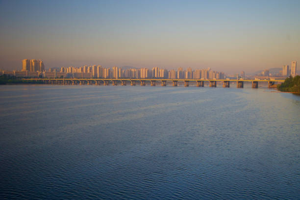 The Han River in Seoul welcomes the day with a tranquil dawn, mirroring the soft hues of the morning sky. This serene scene captures the quietude of the river, offering a moment of peace in the bustling heart of the city, a testament to the natural beauty amidst urbanity.