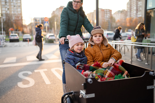 Mother riding her little daughter and son in cargo bike on parking lot in front of shopping mall on cold winter day