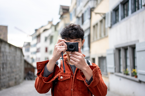 Asian backpacker use camera taking a picture of beautiful landscape. Young male tourist traveler travel alone on street in city, using cellphone record vlog on holiday vacation trip in Switzerland.