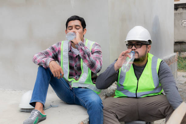 dos trabajadores de la construcción asiáticos con chaleco y casco de seguridad, cansados, sedientos, hidratados, agua potable de botellas de plástico, relajados, sentados en el sitio de construcción en un día soleado sobrecalentado - desempleo construccion fotografías e imágenes de stock