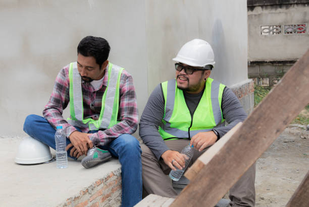 istock Two Asian or Indian builder workers wearing safety reflected vests and helmets tired thirsty hydrated drinking water from plastic bottles together relaxed sitting on construction site talking together 1840139448