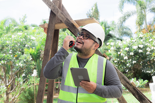 Portrait young overweight Asian engineering architect foreman male wearing safety vest and helmet using walkie-talkie radio on construction site, contractor modern working holding tablet technology