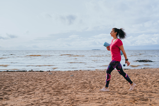 A fit Latin American girl in sportswear sneakers carries a sports mat in her hands walks along the beach going to work out meditate and practice yoga against view in ocean. Outdoor exercise.