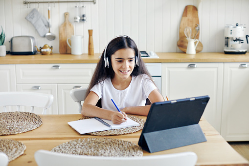 Pretty pre-teen 12s girl in wireless headphones sit at table e-learning, listen on-line course, audio lesson, receive new knowledge, skills using internet and modern tech. Child development. Education