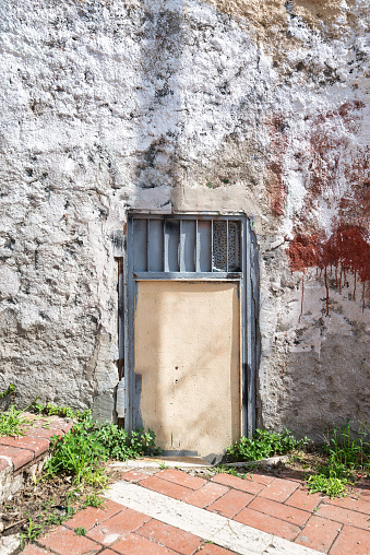 Weathered metal door set within grunge stone wall in an abandoned resedential area. The surrounding stone wall is rough-hewn, creating an atmosphere of mystery and intrigue