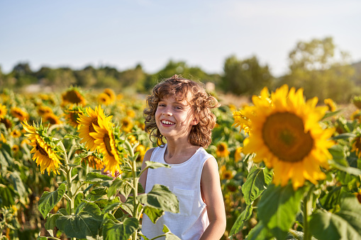 Portrait of smiling preteen male in white dress looking at camera while standing amidst sunflower plants in countryside