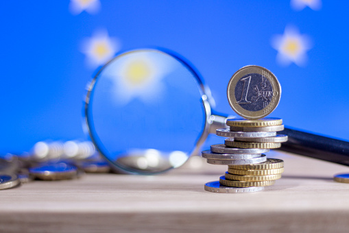 Magnifying glass standing next to unstable stack of coins with one euro coin on top and flag of European Union in background illustrating investigating finance in EU or looking for money laundering or counterfeited money.
