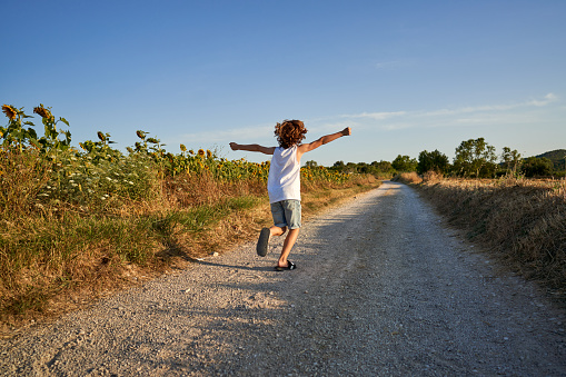 Back view of anonymous child with flying curly hair spreading arms while running along rural road near sunflower field on summer day