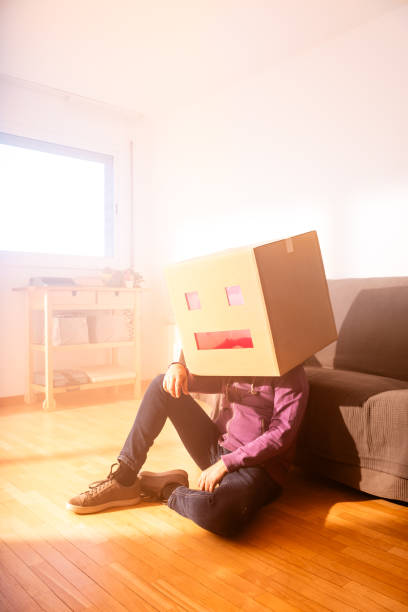 man with robot face box on head sitting near sofa - solitude loneliness hardwood floor box imagens e fotografias de stock