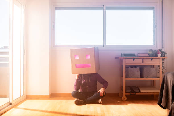 Full body of faceless male sitting on parquet floor leaning on a wall while hiding head in cardboard box with square eyes and rectangular mouth on sunny day