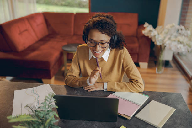 istock Smiling female student in headphones studying remotely on laptop from home and making notes 1840360384
